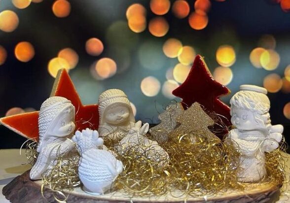 Presepe in miniatura adagiato su un tronco di legno, decorato con paglia oro, alberi e stelle di ceramica dipinta di rosso, per un’atmosfera natalizia calda e incantata.